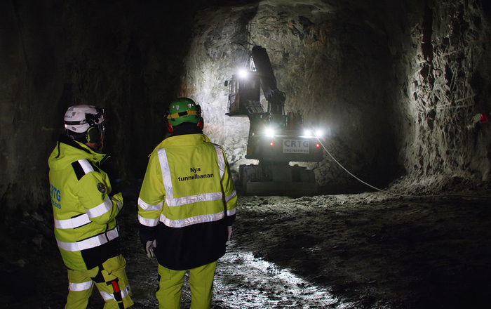 Två personer i arbetskläder med olika företagsnamn, står i en tunnel. Ser på en arbetsmaskin som arbetar med en bergvägg längre bort.
