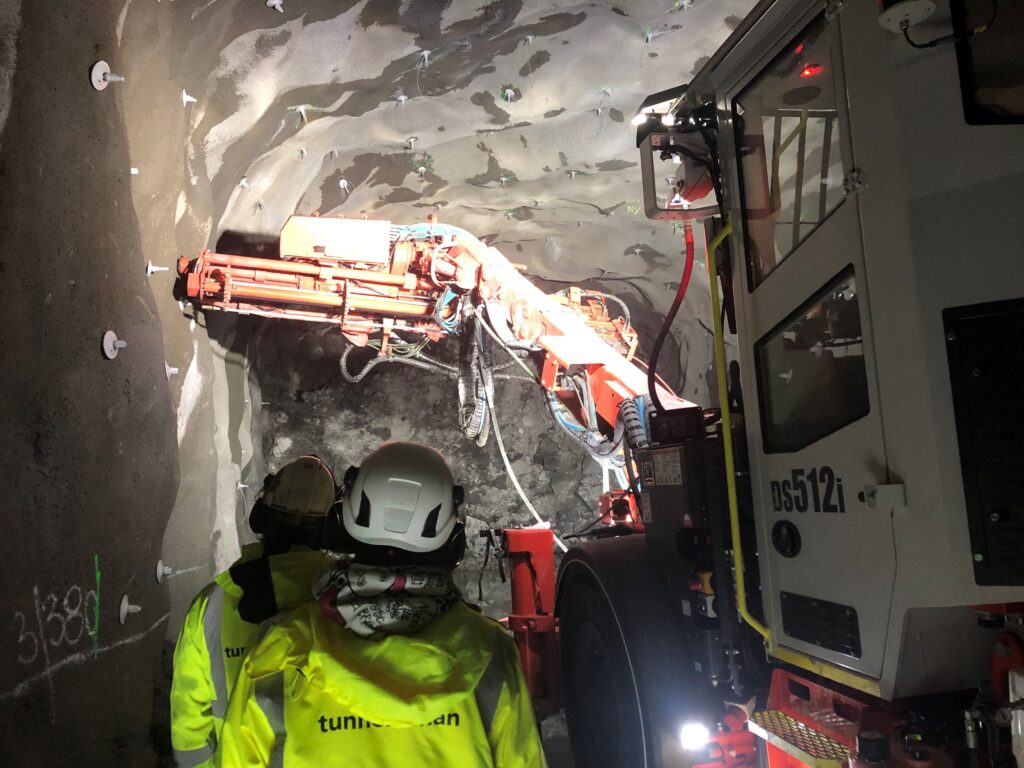 Bultmaskin jobbar. Två arbetare i gula jackor tittar på arbetet. 