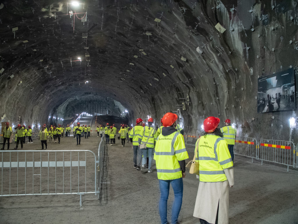 Människor med gula västar och röda hjälmar går omkring i en stor tunnel.