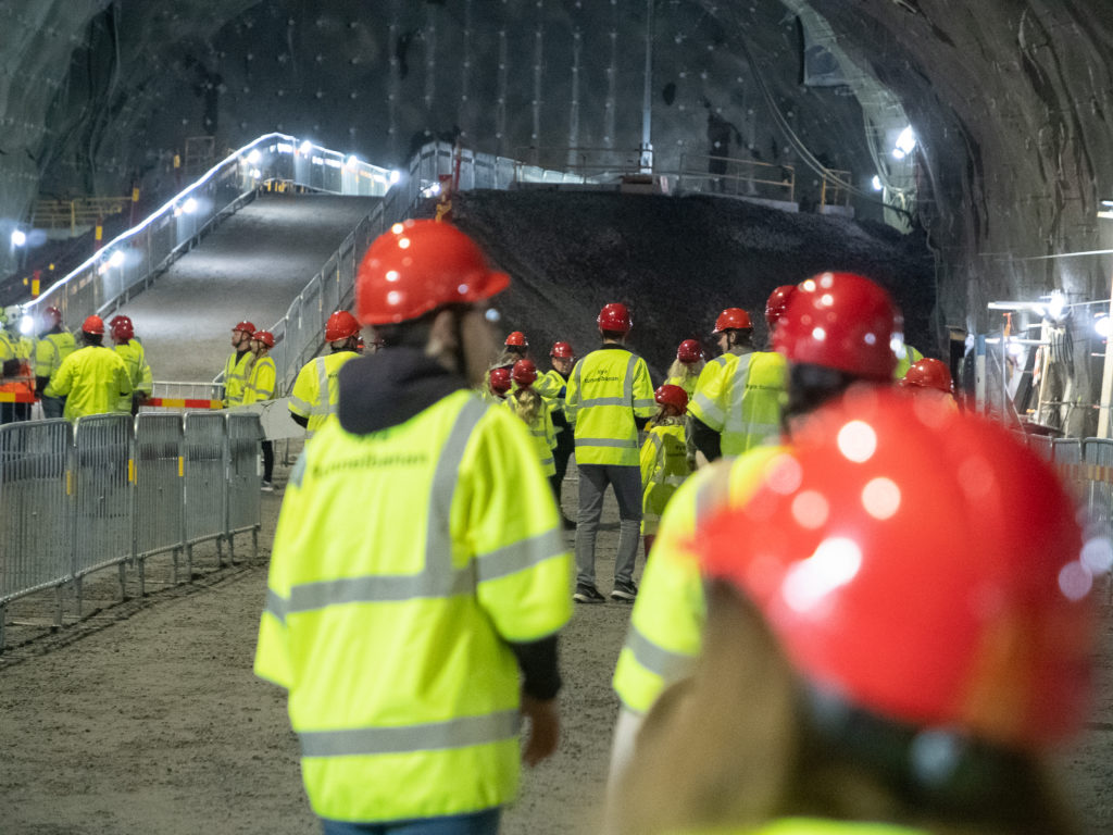 Människor med gula västar och röda hjälmar går omkring i en stor tunnel.