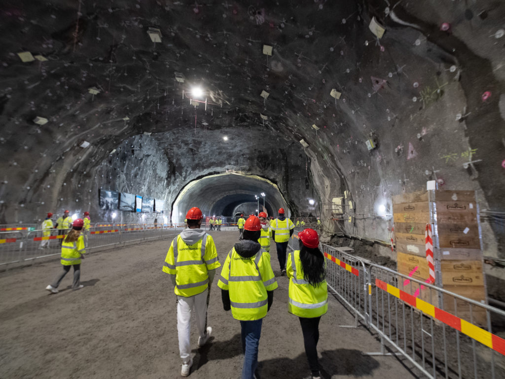 Människor med gula västar och röda hjälmar går omkring i en stor tunnel.