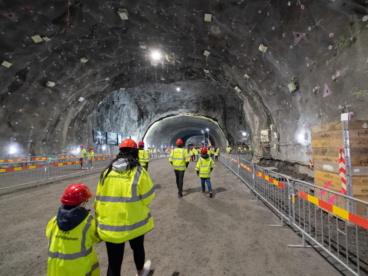 En stor tunnel. Människor går med i tunneln med gula västar och röda hjälmar.
