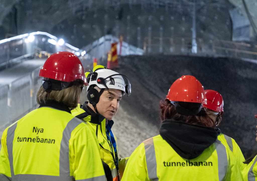 En man i vit hjälm står och pratar med en grupp människor i röda hjälmar.