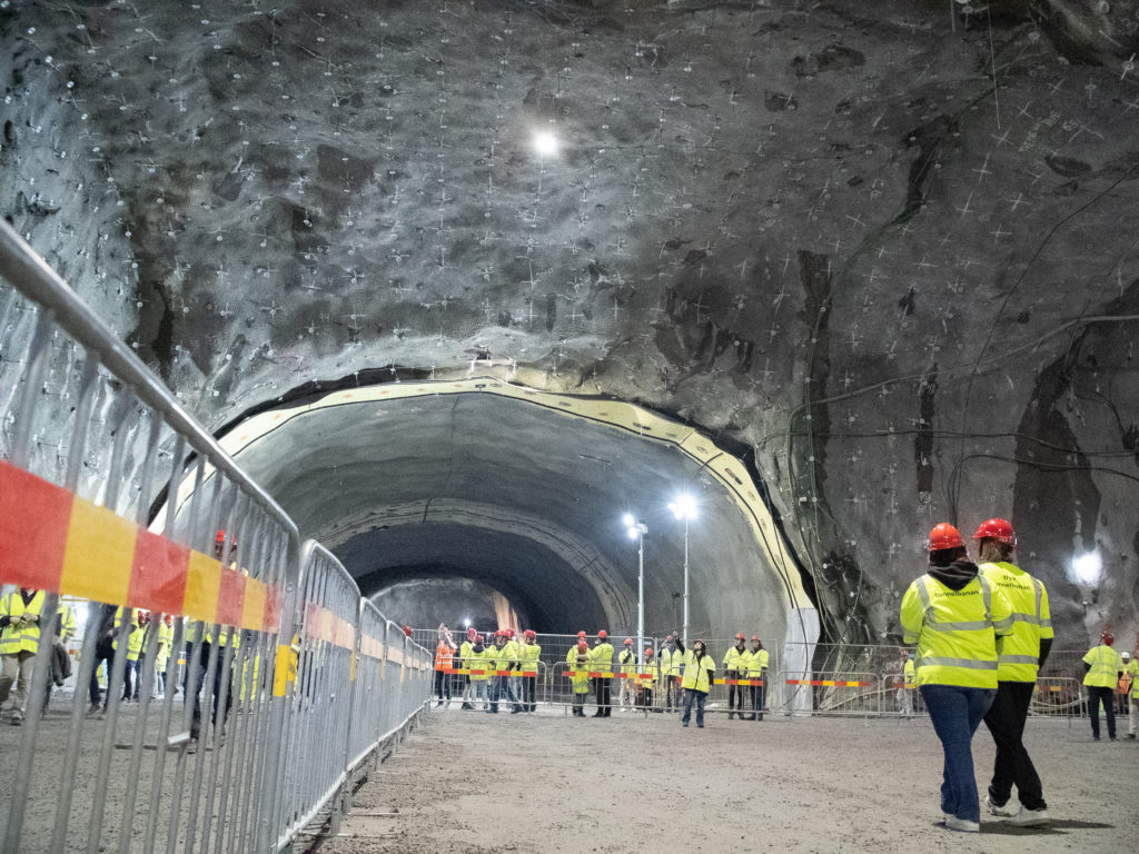 Människor med gula västar och röda hjälmar går omkring i en stor tunnel.