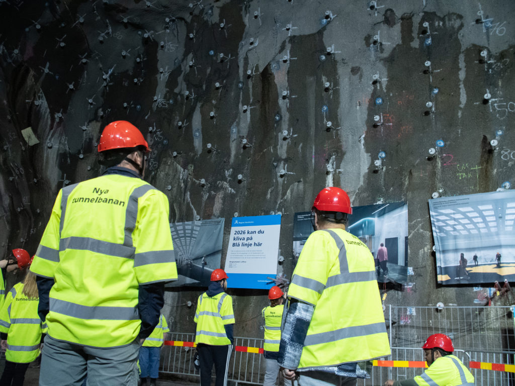 Människor med gula västar och röda hjälmar går omkring i en stor tunnel. De tittar på skyltar som visar visionsbilder av den nya tunnelbanestationen.
