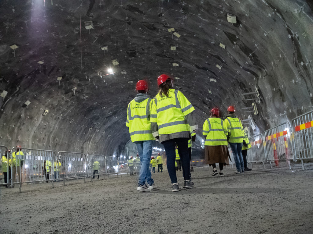 Människor med gula västar och röda hjälmar går omkring i en stor tunnel.
