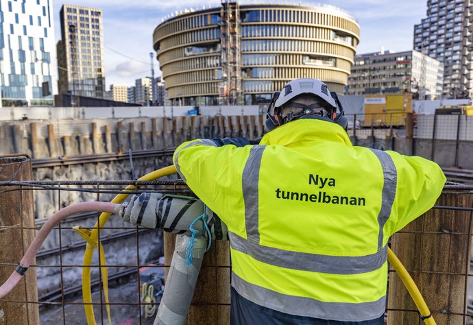 Arbetare i neongul jacka med texten "Nya tunnelbanan" på ryggen står vid en byggarbetsplats. Stadsbebyggelse och ett runt höghus syns i bakgrunden.