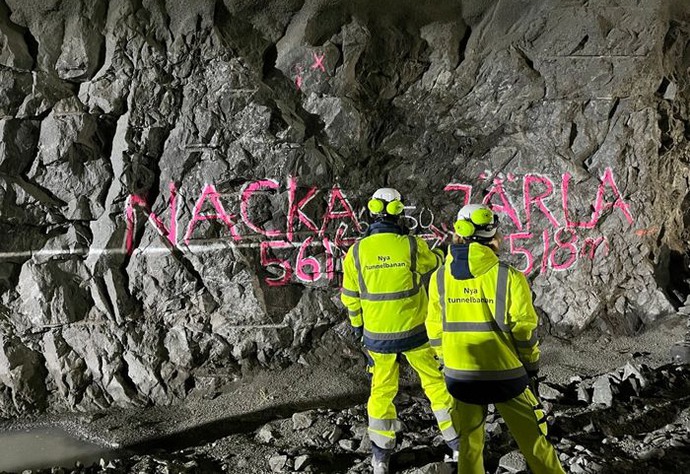 Två personer i gula kläder står framför en bergvägg. På väggen står det "Nacka 561 m" och en pil åt vänster samt "Järla 518 m" och en pil åt höger.